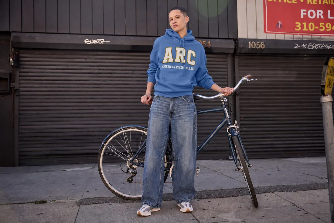 Student in a college-branded blue hoodie standing with a bike on a city sidewalk